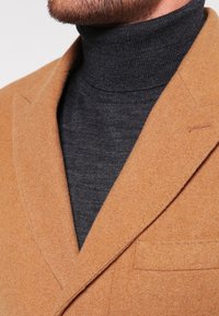 Blazer de lana color camello con solapas, combinado con un suéter de cuello alto en gris oscuro. El blazer presenta una textura suave y un bolsillo en el pecho.