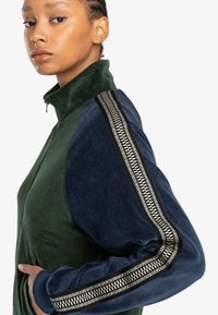 Chaqueta verde con cremallera y mangas de terciopelo azul oscuro, que presenta un patrón de rayas decorativas a lo largo de los brazos y bolsillos laterales.