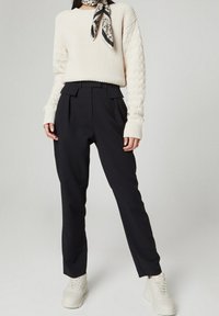 Roomkleurige geribbelde trui met gestructureerde mouwen, gecombineerd met donkere, hoog opgesneden broek en witte sneakers. Een sjaal met patroon maakt de outfit af.