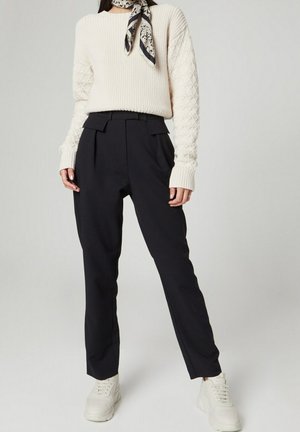 Krämfärgad ribbstickad tröja med texturerade ärmar, kombinerad med mörka byxor med hög midja och vita sneakers. En mönstrad scarf kompletterar outfiten.