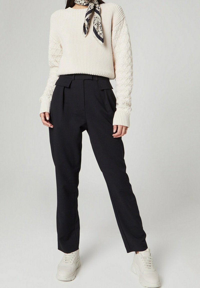 Cremefarbener, gerippter Pullover mit strukturierten Ärmeln, kombiniert mit dunkler, hochtaillierter Hose und weißen Sneakers. Ein gemustertes Halstuch setzt einen Akzent im Outfit.