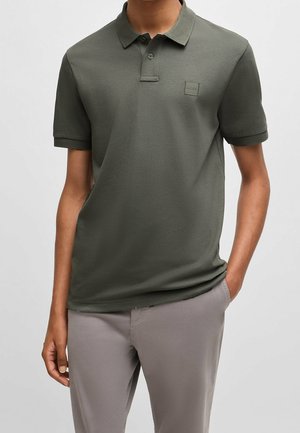 Mann trägt olivgrünes kurzärmeliges Poloshirt mit Knöpfen und hellgraue Hose, Hand in der Tasche, neutraler Hintergrund.