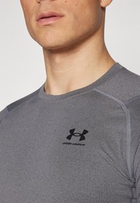 Graues Sportshirt aus weichem Material. Mit rundem Halsausschnitt und kontrastierenden Nähten. Schwarzes Under Armour-Logo auf der Brust.