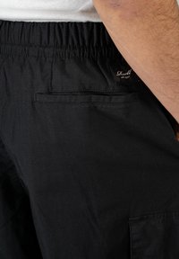 Schwarze Cargo-Hosen mit elastischem Bund, ausgestattet mit einer Gesäßtasche und einem kleinen Markenlabel. Texturiertes Gewebe mit feinen Linien.