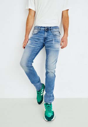 Homme portant un jean bleu clair, un t-shirt blanc et des baskets vertes Adidas, debout contre un mur blanc avec la jambe légèrement pliée.