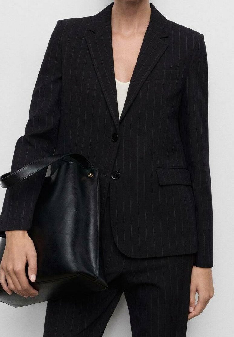 Personne portant un costume noir à fines rayures tenant un grand sac cabas en cuir noir avec un bouton-pression doré.