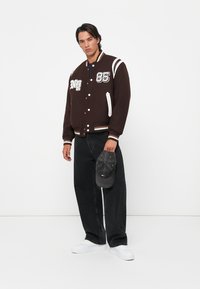 Giacca varsity marrone con accenti bianchi e numero "85," abbinata a pantaloni larghi neri e sneakers bianche. Tiene in mano un cappellino grigio.