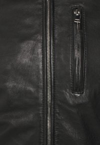 Zwarte leren jas met zilveren ritsdetails. Beschikt over een ritszak aan de linkerkant en heeft een gladde, gestructureerde afwerking.