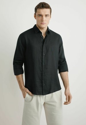 Mężczyzna o jasnej karnacji i krótkich brązowych włosach nosi czarną bluzkę z długim rękawem i jasnobeżowe spodnie, stoi z jedną ręką w kieszeni.