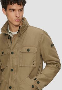 Beige jas met een hoge kraag, meerdere zakken, zwarte knopen en verstelbare manchetten. Gemaakt van een lichte stof met een gladde textuur.