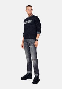 Suéter de punto gris oscuro con el logo "GUESS", combinado con jeans gris desgastados y botas negras con cordones. Presenta puños y dobladillo a rayas.