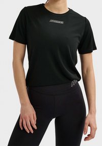 Černé sportovní tričko s krátkým rukávem, s nenápadným bílým logem značky přes hruď. Hladká, lehká tkanina.