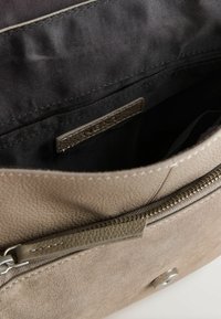 Nahast käekott taupe toonis, millel on tekstuuriga pind, lukk ja musta kangaga vooder, näidates sisemist tasku silti.