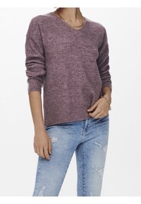 Personne portant un pull violet à col en V côtelé avec les manches retroussées et un jean bleu clair déchiré, accessoirisé de bijoux en or.
