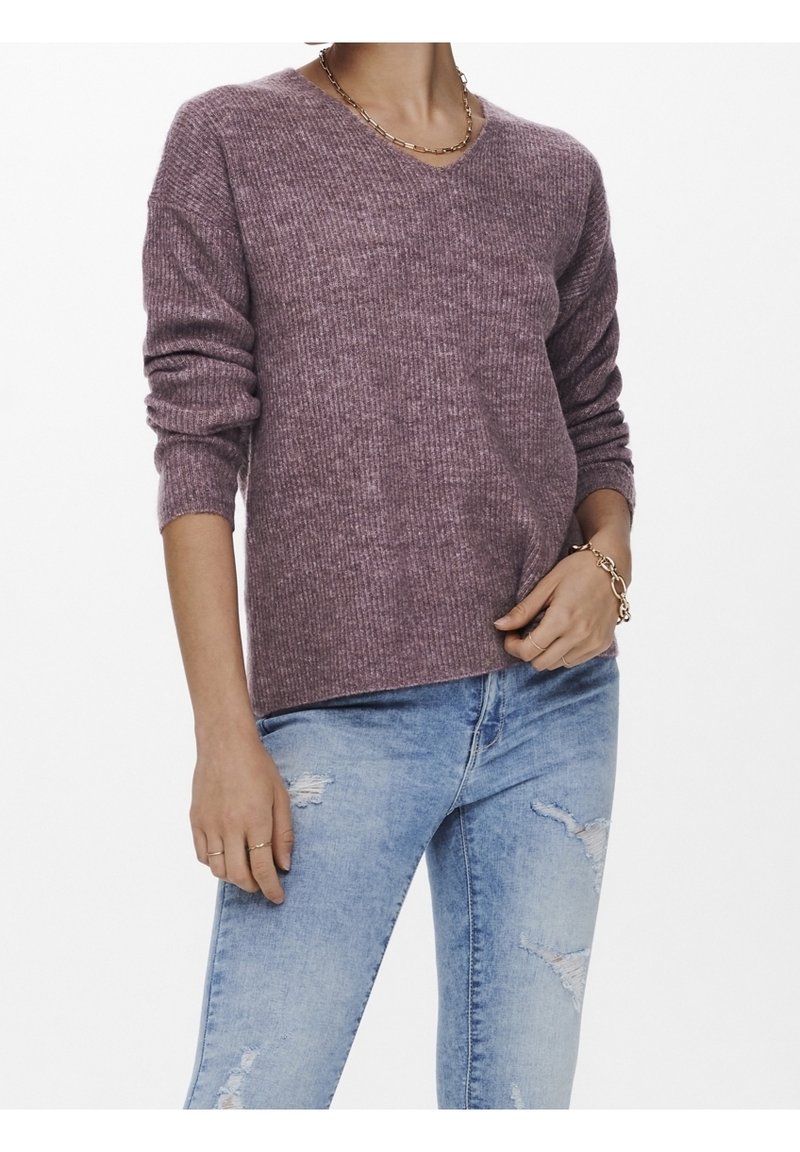 Personne portant un pull violet à col en V côtelé avec les manches retroussées et un jean bleu clair déchiré, accessoirisé de bijoux en or.