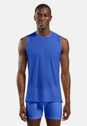 Mann steht frontorientiert und trägt ein ärmelloses blaues Sportshirt sowie passende enganliegende Shorts vor einem schlichten Hintergrund.