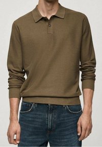 Pitkähihainen polo, joka on valmistettu tekstuurisesta oliivinvihreästä kankaasta, jossa on klassinen kaulus ja kolmesta napista koostuva nappilista, yhdistettynä tummansinisiin farkkuihin.