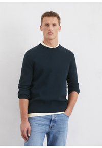 Marineblauer, gerippter Pullover mit Rundhalsausschnitt, langen Ärmeln und einem weißen Kragen darunter, kombiniert mit hellblauen Jeans.