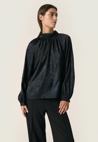 Blusa nera con collo alto, dettagli arricciati e maniche lunghe. Realizzata in un tessuto lucido e testurizzato, abbinata a pantaloni neri.