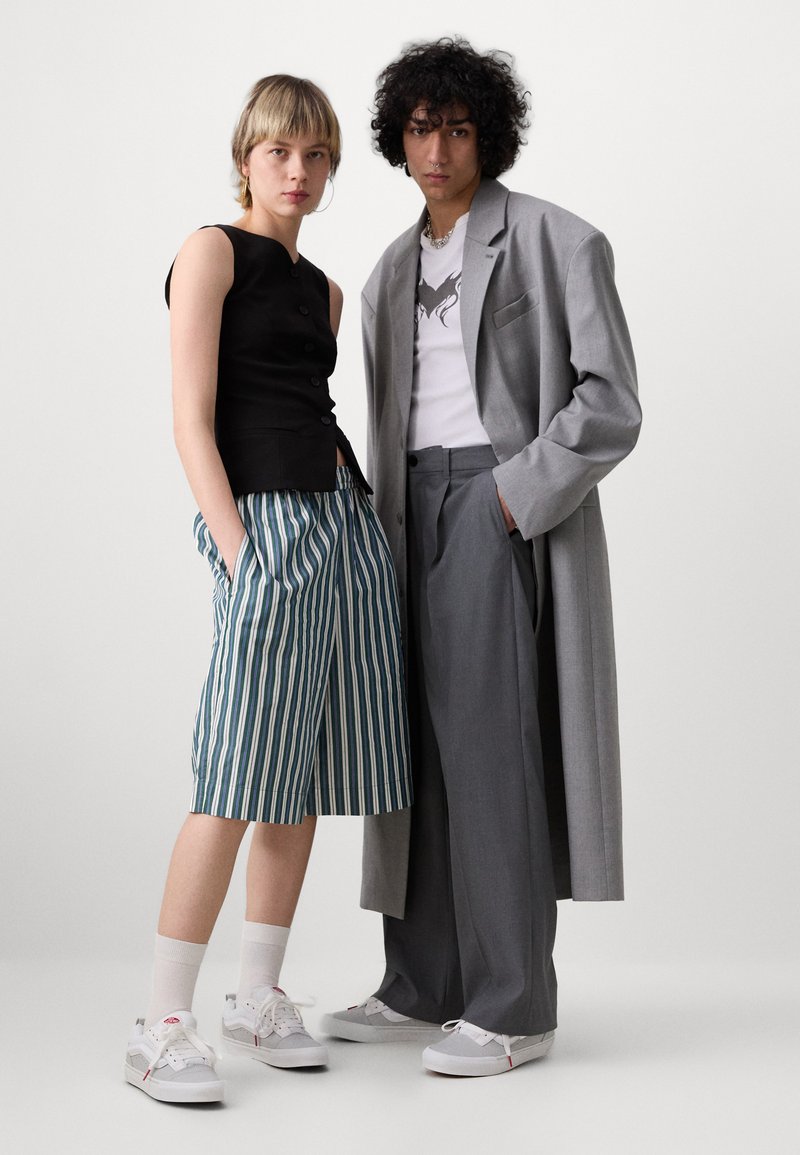Chaleco negro con botones, pantalones cortos de rayas azul y blanco, abrigo gris de corte amplio, zapatillas blancas y calcetines neutros. Tejido texturizado, ajuste entallado.