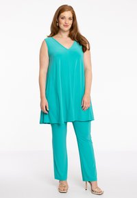 Turquoise mouwloze tuniek met V-hals en een vloeiende snit, gecombineerd met bijpassende strakke broek. Vloeiend, lichtgewicht stof met een subtiele glans.
