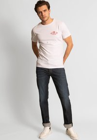Vit t-shirt med kort ärm och röd "rePLAY"-logotyp, i kombination med mörkblå jeans och ljusa sneakers. Enkelt, figurnära snitt.