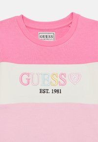 Rosa Rundhalsausschnitt T-Shirt mit weißem Querstreifen. Verfügt über gesticktes "GUESS" in mehreren Farben und "EST. 1981" in Schwarz. Weicher Baumwollstoff.