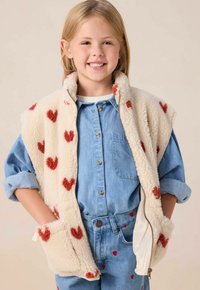 Fleece vest in cream with red heart patterns; zip closure and pockets. Worn over a blue denim shirt, paired with coordinated heart-embroidered jeans.