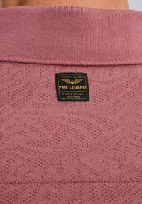 Polo-shirt en tricot rose avec des motifs texturés, comportant une étiquette noire rectangulaire indiquant "PME LEGEND" à la nuque arrière.
