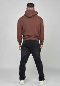 Felpa marrone con un grande cappuccio, abbinata a jeans neri. Dettagli di cucitura visibili, polsini a coste e design minimalista. Sneakers bianche.