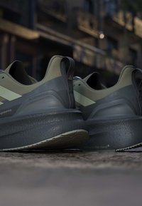 Ein Paar graue und olivgrüne Sportschuhe, die im Freien auf einer groben Oberfläche mit unscharfem städtischem Hintergrund platziert sind.