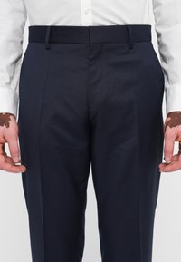 Pantaloni de stofă bleumarin, croiți, cu fața plată, buzunare laterale și curea, confecționați dintr-un material neted, asortați cu o cămașă albă cu butoni.