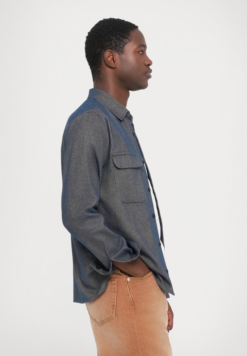 Camicia da uomo button-up in tessuto grigio e blu, con due taschini sul petto, maniche lunghe e colletto; indossata con pantaloni marroni chiari.