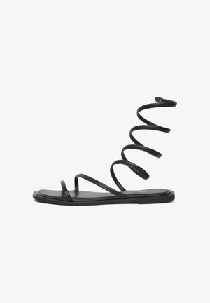 Schwarze flache Sandale mit dünnen Riemen und einem langen spiralförmigen Riemen, der um Knöchel und Unterschenkel gewickelt werden kann.
