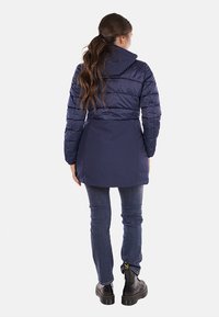 Giacca puffer blu navy con cappuccio, caratterizzata da un design aderente, texture trapuntata e vita cintrata, abbinata a jeans blu e robusti stivali neri.