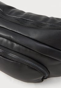 Bolsa de couro preta com dois zíperes visíveis e texto em relevo sutil sobre um fundo branco limpo.