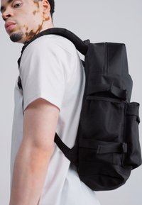 Mochila negra con un diseño estructurado, detalles de malla, correas ajustables y múltiples bolsillos para almacenamiento. Llevada en la espalda de una persona.