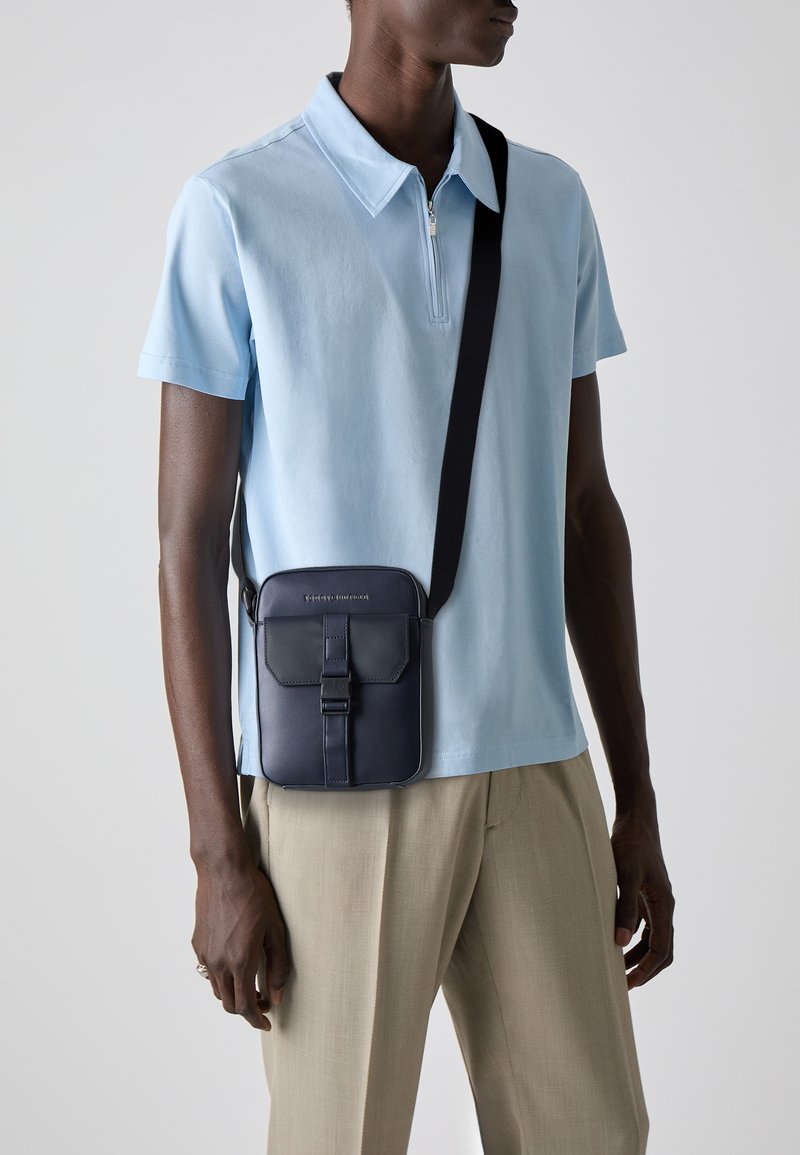 Navy leather crossbody bag with flap closure, adjustable strap, and rectangular shape. Worn over a light blue polo and beige pants.