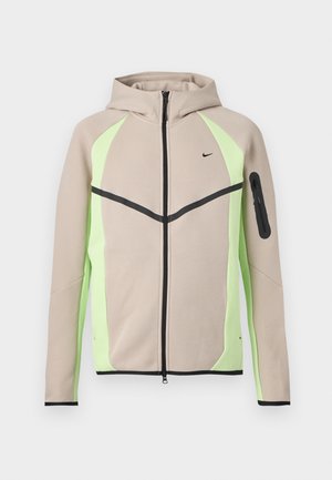 Beige-grüne Kapuzenjacke mit Reißverschluss, schwarzen Akzenten, kleinem Nike-Logo auf der Brust und Reißverschlusstasche am linken Ärmel.
