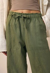 Pantaloni verzi din amestec de in, cu talie elastică, șnur și două buzunare frontale. Au o croială lejeră și o textură fină.