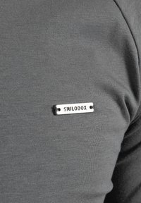 Grijze, gestructureerde stof met een aansluitend ontwerp. Beschikt over een metalen label met de tekst "SMILODOX" op de borst genaaid.