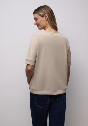 Beige kortærmet top med afslappet pasform, rund halsudskæring og opsmøgede ærmekanter, kombineret med mørkeblå jeans, vist bagfra.