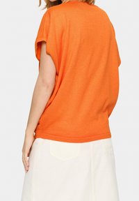 Lyhythihainen oranssi paita rentoa istuvuutta, valmistettu pehmeästä kankaasta. Takakappale on hieman pidempi, yhdistetty valkoisiin hameeseen, jossa on taskut.