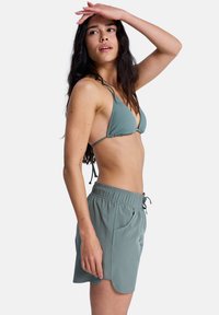 Τοπ μπικίνι τριγωνικό και σορτς με κορδόνι σε απαλό πράσινο ύφασμα. Τα σορτς διαθέτουν ελαστική μέση και τσέπες στα πλάγια. Ομαλή υφή.