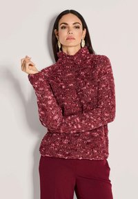 Vrouw met lang donker haar, die grote gouden blaadjesoorbellen draagt, een dikke rode en roze gebreide trui en donkerrode broek, poseert met gekruiste armen.