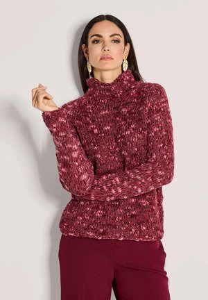 Frau mit langen dunklen Haaren, die große goldene Blätter-Ohrringe, einen dicken rot-pinken Strickpullover und dunkle rote Hosen trägt, posiert mit verschränkten Armen.