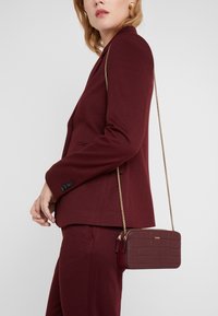 Punainen krokotiilinanatasku, jossa on kultaketju, vetoketjukiinnitys ja suorakulmainen muoto, käytetään viininpunaisen puvun kanssa.