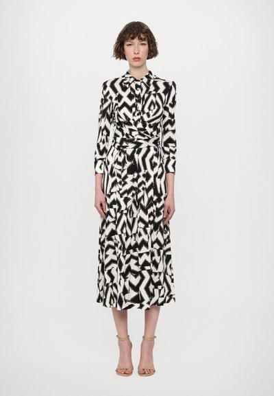 Rochie alb-negru cu model abstract, mâneci lungi, închidere cu nasturi și talie strânsă. Sandale cu curea la gleznă completează aspectul.