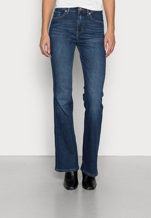 Personne portant un jean bootcut bleu foncé associé à des bottes noires à bout pointu, debout contre un mur clair uni.