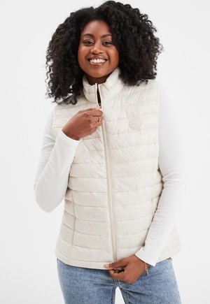 Gilet imbottito color crema con cuciture orizzontali trapuntate, colletto alto e zip frontale. Indossato sopra una maglietta a maniche lunghe bianca.